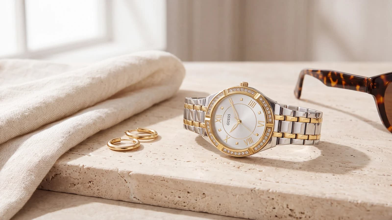 Montre Guess Cosmo bicolore à lunette sertie de cristaux posée sur de la pierre claire avec des créoles dorées